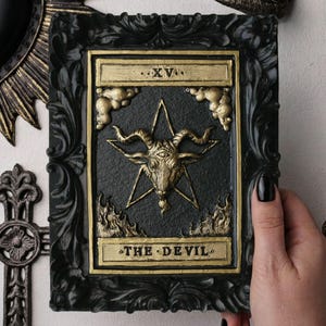 May include: A rectangular wall plaque featuring a relief of The Devil tarot card, XV, with a central goat head and pentagram in gold on a black background, encased in an ornate, textured black frame with gold accents and decorative clouds.