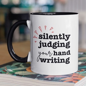 May include: White ceramic mug with a black handle and rim. The mug features the text "silently judging your handwriting" in black and red lettering. The mug is sitting on a book with a floral design.