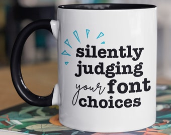 Puede incluir: Taza de cerámica blanca con borde y asa negros. La taza tiene un diseño azul y negro que dice "silently judging your font choices".