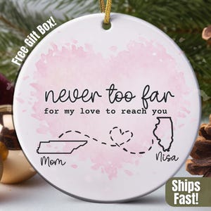 May include: A white ceramic ornament with the text "never too far for my love to reach you." It features a pink watercolor background, outlines of Tennessee and Illinois, and the words "Mom" and "Nisa."