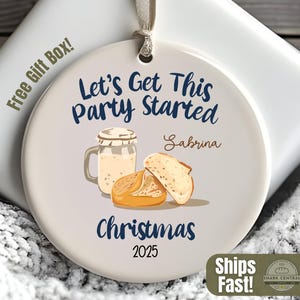 Peut inclure: Ornement de Noël rond en céramique blanche avec le texte « Let's Get This Party Started » et « Christmas 2025 ». Il présente des illustrations d'un bocal et de pain. Un coffret cadeau est en arrière-plan.