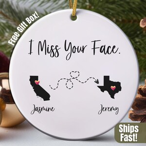 May include: White ceramic ornament with the text "I Miss Your Face." Features black silhouettes of California and Texas, each with a pink heart. Names "Jasmine" and "Jeremy" are below the states. A gold string is attached for hanging.