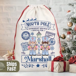 May include: A cream Christmas sack with a red drawstring and trim. It features a North Pole design with nutcracker illustrations and the name "Marshall". Text includes "Overnight Service" and "Do Not Open Until Dec 25".
