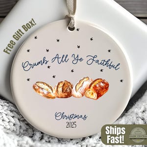 May include: A circular Christmas ornament with the words "Crumb All Ye Faithful" and "Christmas 2025". It displays a watercolour illustration of four loaves of bread. The ornament is on a white gift box.