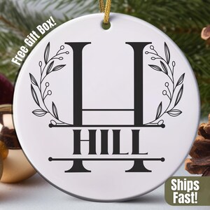 May include: White ceramic Christmas ornament with a gold string, featuring a black monogram "H" above the name "HILL". The design includes floral accents and is set against a festive background with ornaments and pine cones.