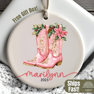 Puede incluir: Adorno redondo color crema con una ilustración en acuarela de botas de vaquero rosas adornadas con arreglos florales. El adorno tiene el nombre "marilynn" y el año "2025" escrito. El texto "Free Gift Box!" también es visible.