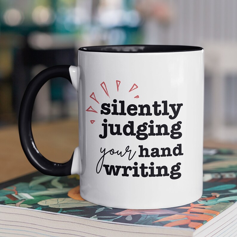 May include: White ceramic mug with a black handle and rim. The mug features the text "silently judging your handwriting" in black and red lettering. The mug is sitting on a book with a floral design.