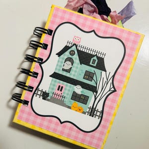 May include: A spiral-bound Halloween-themed journal with a pink gingham cover and a yellow border. The cover features a haunted house illustration with ghosts, pumpkins, and a black cat. Decorative ribbons in pink, purple, and black are attached to the top.