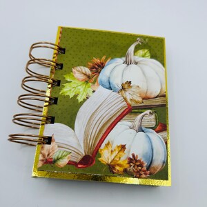 May include: A small spiral-bound notebook with a gold metallic edge. The cover features a green background with a pattern of white pumpkins, an open book, and colorful autumn leaves. The binding consists of five bronze-colored metal wire loops.