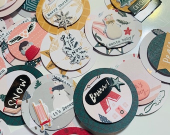 May include: A collection of 12 round paper craft embellishments with winter holiday themes. The embellishments feature various designs, including snowmen, snowflakes, trees, and houses. The embellishments are decorated with gold, red, green, and white colors. The text on the embellishments includes "Let it snow", "Merry Christmas", "Catching snow", "Santa baby", "Brrr", "It's December", "Nice & Cozy", "Hey snow", "Silent night", "Joy", "Peace", and "Wonderful".