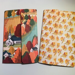 May include: Two notebooks with different designs. The notebook on the left has a colorful illustration of a church and houses in a rural setting. The notebook on the right has a white background with a repeating pattern of yellow and orange flowers.