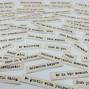 May include: A collection of small, rectangular tags with inspirational phrases printed in a typewriter font. Phrases include "stay positive," "miracles happen," and "enjoy the journey." The tags are beige with black text.