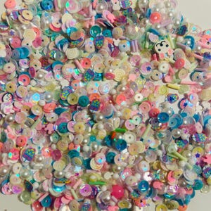 May include: A colorful assortment of craft supplies, including sequins, beads, and small decorative pieces. The mix features various shapes, sizes, and colors, such as pink, blue, white, and yellow. Some pieces have a pearlescent sheen, and there are small bunny and candy-shaped embellishments.