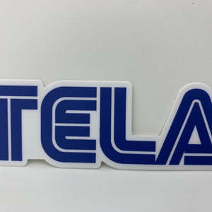 May include: A white sticker with the word "TELA" in a bold, stylized font. The letters are blue with a white outline, creating a layered effect. The sticker has a clean, modern design.