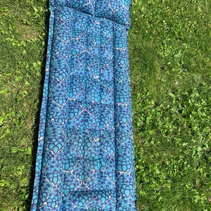 Puede incluir: Una colchoneta para exteriores con estampado azul y turquesa con una almohada adjunta. La colchoneta es rectangular y presenta un diseño floral repetido. La colchoneta está sobre un lecho de hierba verde.