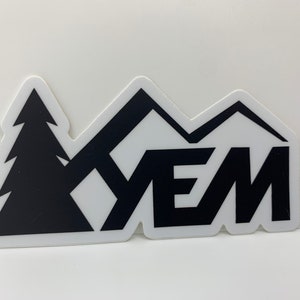 May include: Black and white sticker with a stylized mountain range and a pine tree. The word "YEM" is written in bold letters.