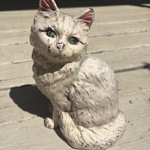 May include: A vintage, off-white cat figurine with green eyes and pink-lined ears. The cat is sitting upright, with a fluffy coat and a slightly distressed, aged appearance. The figurine is made of a solid material.