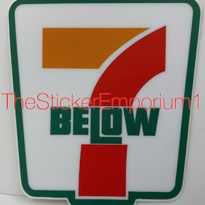 May include: A white sticker with a green border featuring a stylized "7" logo in orange and red, with the word "BELOW" in green block letters. The sticker has a rounded rectangular shape.