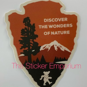 May include: An orange and brown sticker with a silhouette of a mountain, a tree, and a bear. The sticker has the text "Discover the Wonders of Nature".