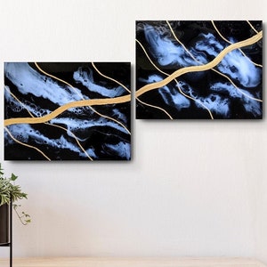 Puede incluir: Dos pinturas abstractas con acentos negros, blancos y dorados. Las pinturas presentan patrones azules y blancos en espiral con finas líneas doradas. Las pinturas se exhiben en una pared blanca sobre un estante de madera.