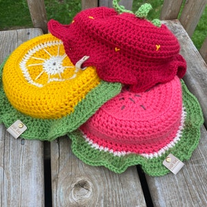 May include: Three crocheted fruit-shaped baskets, a red strawberry, a yellow orange, and a pink watermelon, with green crocheted leaves and white trim. Each basket has a small brown leather tag with the text "High End Handmade" on it.