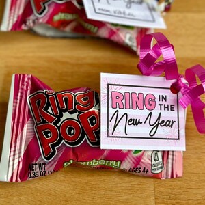 May include: A pink and white gift tag with the text "Ring in the New Year" attached to a pink and white Ring Pop candy wrapper with a pink ribbon.