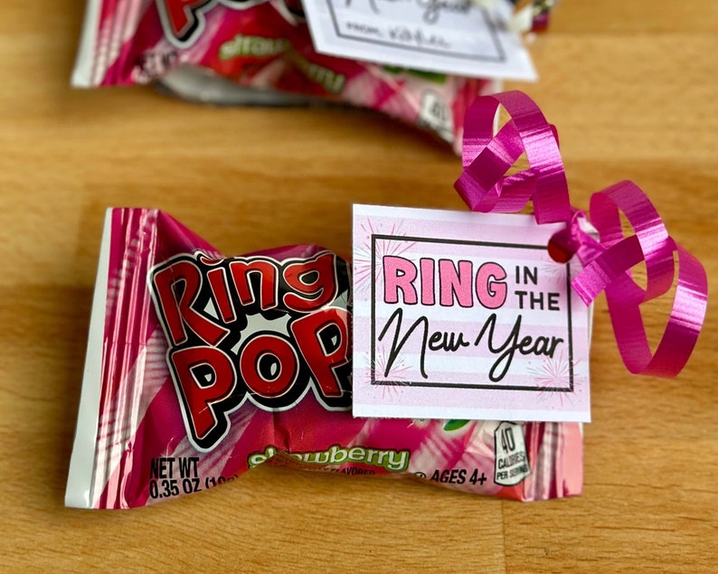May include: A pink and white gift tag with the text "Ring in the New Year" attached to a pink and white Ring Pop candy wrapper with a pink ribbon.