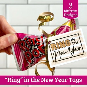 May include: A printable New Year's Eve gift tag with the text "Ring in the New Year" in gold lettering on a striped background. The tag is tied to a pink and white Ring Pop candy with gold ribbon.
