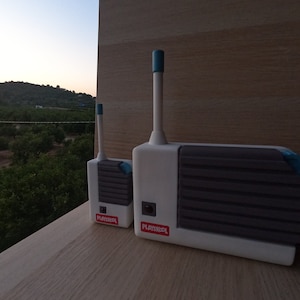 May include: Two white and gray toy walkie-talkies with blue accents and antennas. The larger one has the word "Playskool" on it. The background shows a view of trees and a mountain.