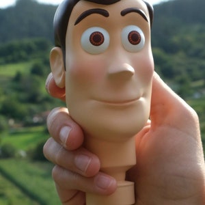 May include: A Woody figure head, featuring a tan complexion, brown hair, and large, expressive eyes. The figure is held in a hand against a blurred green background, evoking a sense of nostalgia and play.