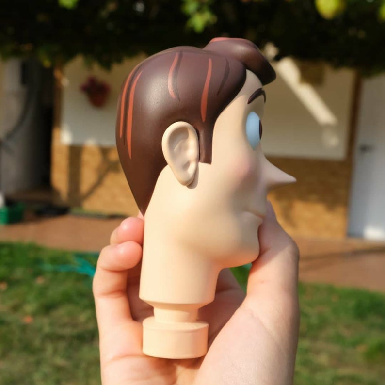 May include: A figurine head with brown hair, a tan face, and blue eyes. The head has a prominent nose and is held in a hand. The figurine is a cartoon character.