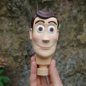 May include: A detailed, three-dimensional figure of a cartoon character's head. The figure has brown hair, large eyes with brown irises, and a smiling expression. The skin tone is light, with rosy cheeks. The figure is held in a hand.