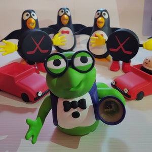 Puede incluir: Colección de figuras caprichosas. Se ve una rana verde con gafas y un pingüino con un disco de hockey. También hay coches de juguete rojos. La rana lleva un esmoquin. Los pingüinos tienen manos y pies amarillos.