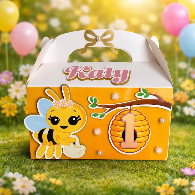 May include: A white and orange party favor box with a bee theme. The box features a cartoon bee, a honeycomb with the number 1, and the name "Katy". The box is decorated with a gold bow and pearls.