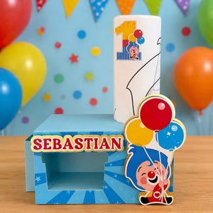 May include: A blue box with the name "SEBASTIAN" in red and gold lettering, a white candle with a "1" and cartoon character, and a cartoon character holding balloons. Colorful balloons and a banner are in the background.