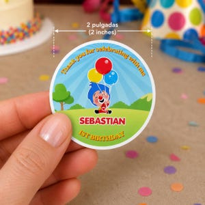 May include: A round sticker, 2 inches in diameter, featuring a cartoon clown holding balloons. The sticker has the text "Thank you for celebrating with me," "Sebastian," and "1st Birthday." The design includes a blue sky, green grass, and a small tree.
