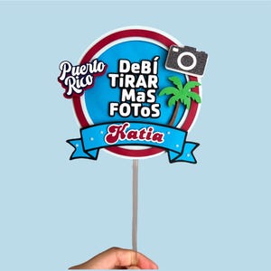 May include: A round cake topper with a blue and red border, featuring the text "Puerto Rico" and "Debí Tirar Más Fotos Katia". Includes a camera and palm tree graphic. Held by a hand against a light blue background.