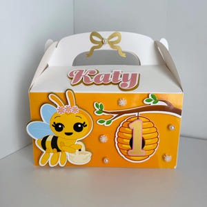 May include: A white and orange party favor box with a bee theme. The box features a cartoon bee, a honeycomb with the number 1, and the name "Katy". The box has a handle and a gold bow.