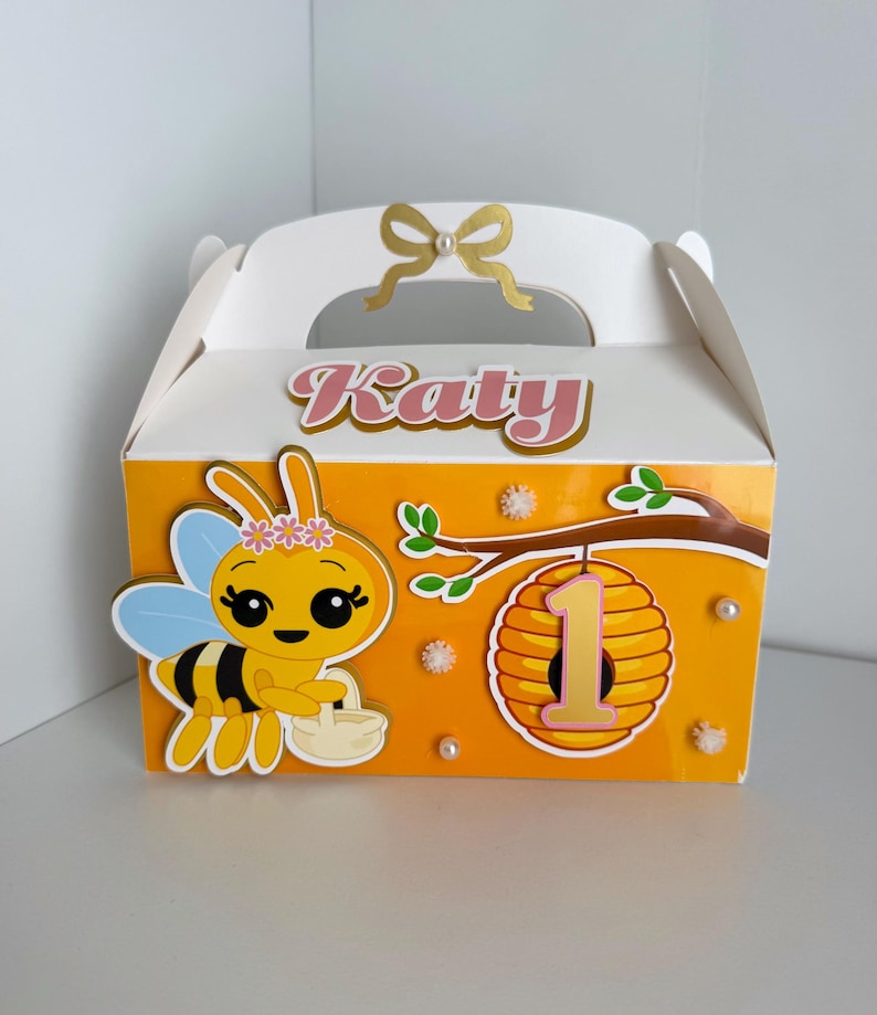May include: A white and orange party favor box with a bee theme. The box features a cartoon bee, a honeycomb with the number 1, and the name "Katy". The box has a handle and a gold bow.