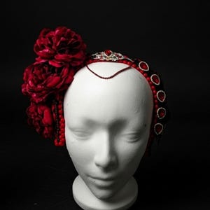 May include: A decorative headpiece featuring large, deep red fabric flowers and a beaded headband with red and black accents. The headpiece is displayed on a white mannequin head against a black background.