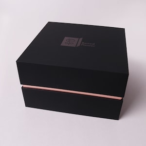May include: A black box with a rose logo and the text "Boxed Flower" on the top. The box has a thin pink stripe around the edge.