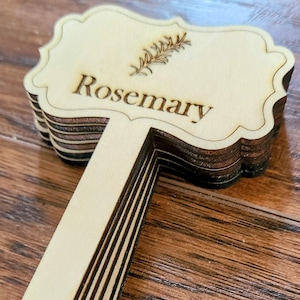 Op de afbeelding: Een stapel houten plantenlabels met een decoratieve, gegolfde rand. Het bovenste label is gegraveerd met het woord "Rozemarijn" en een klein beeld van een rozemarijntak.