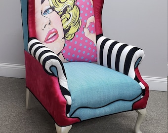 Könnte beinhalten: Ein Ohrensessel mit einem Pop-Art-Design, das ein Frauen Gesicht auf der Rückenlehne zeigt. Der Sessel hat eine rosa, blaue und schwarz-weiß gestreifte Stoffpolsterung. Der Sessel hat silberne Beine.