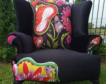 May include: A black wingback armchair with a floral patterned cushion. The cushion features a psychedelic design with mushrooms, flowers, and eyes. The chair has black legs and is sitting on green grass.