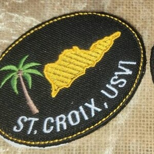May include: Two black embroidered patches with white stitching. One patch features a yellow outline of St. Croix, USVI with a palm tree. The other patch features a white outline of St. Croix, USVI with a red hibiscus flower.