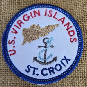 May include: A white embroidered patch with a blue border. The patch features a red and white embroidered text that reads "U.S. VIRGIN ISLANDS" and a blue and white embroidered text that reads "ST. CROIX". The patch also features a brown embroidered outline of an island and a grey embroidered anchor.