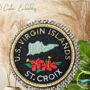 May include: A round embroidered patch with a black background and gold trim. The patch features the text "U.S. VIRGIN ISLANDS" in a gold font, a green outline of the Virgin Islands, and the text "ST. CROIX" in a gold font. The patch also features two red hibiscus flowers.