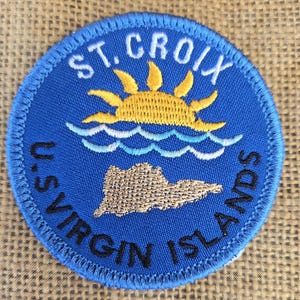 May include: A blue embroidered patch featuring a yellow sun rising over a white wave and a brown island. The text "ST. CROIX U.S. VIRGIN ISLANDS" is written around the edge of the patch.