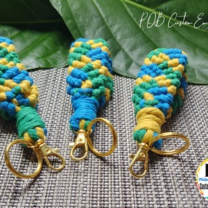 May include: Three colorful keychains with gold key rings. The keychains are made of woven yarn in shades of blue, green, and yellow. The keychains are arranged on a gray textured surface with a green leaf in the background. The text "POB Custom Embroidery" is visible in the top right corner of the image.