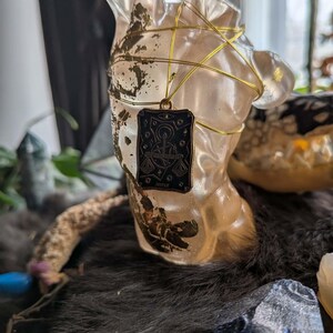 May include: A clear resin bust with gold accents and a gold wire wrap. A black pendant with a gold border hangs from the bust. The pendant features a scale design and the word "JUSTICE".